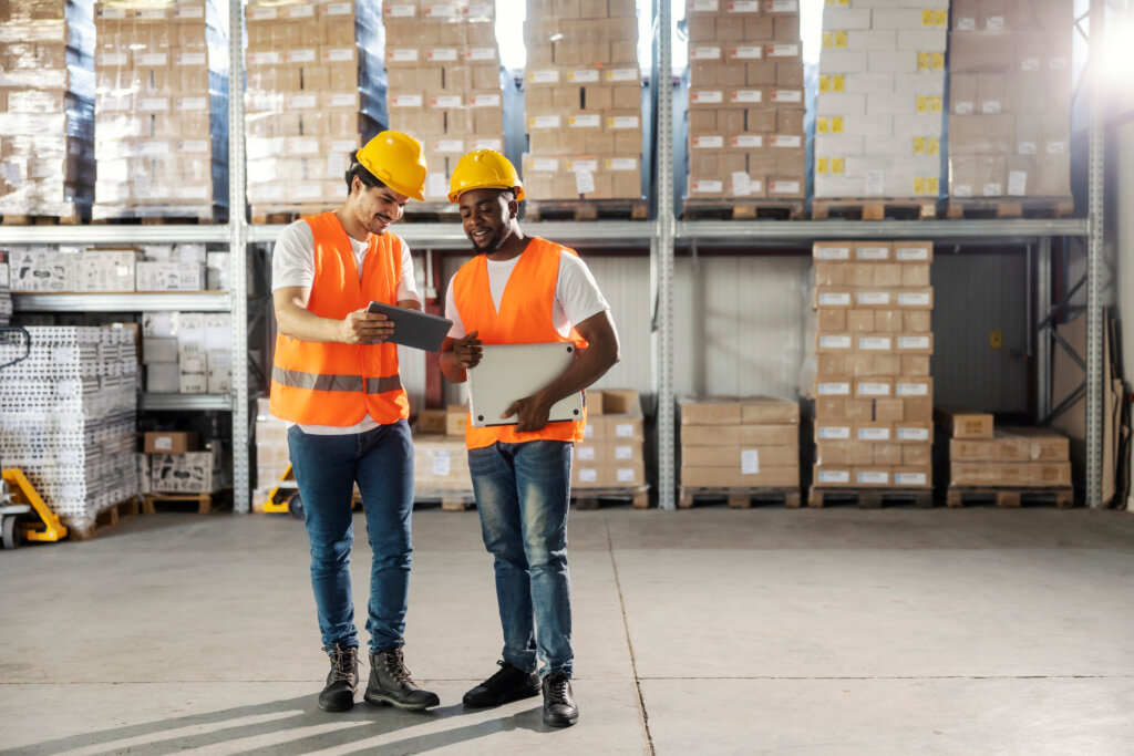 Two multicultural foreign blue collar workers standing at storage with tablet and smiling at it.